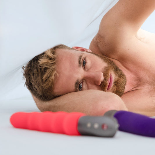 Man laying in bed underneath white sheets with Fun Factory toys
