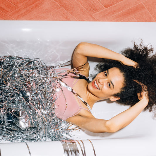 Woman with hand reaching up into curly hair and smiling at the camera, in a bathrub filled with shiny tinsel 